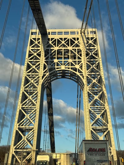 An upward-angled view captures the imposing structure of a massive suspension bridge tower, likely the George Washington Bridge, under a bright, sunny sky. The light-colored steel tower features an intricate lattice of trusses forming X-patterns and a large, prominent arched opening in its upper section. Suspender cables stretch across the frame, disappearing into the sky.

The backdrop is a vibrant blue sky dotted with scattered, fluffy white clouds. Strong sunlight illuminates the bridge, creating sharp contrasts and deep shadows that highlight the geometric complexity of the steel framework.

On the right side of the image, a large white truck, identifiable as a trailer unit, is visible, driving on the bridge deck. Its reflective surface shows hints of the surrounding environment. Clearly visible text on the side of the truck reads "RAVEN TRUCK LEASING, INC.", "1-800-44-SALEM", and "IDEALEASE". Below the main bridge structure, a reflective surface, possibly a lower deck or sound barrier, creates a distorted mirror image of the bridge elements and the sky, adding depth and a sense of vastness to the scene. The overall impression is one of immense architectural scale and robust engineering against a clear, daytime sky.