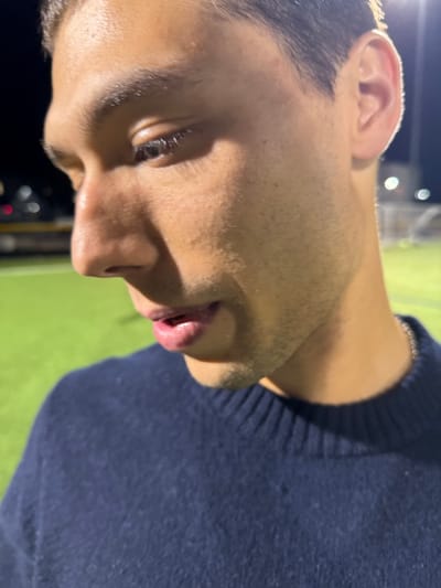 A close-up, angled shot captures the right profile of a young adult male, positioned on an artificial turf athletic field at night in Newton, United States. The individual exhibits a medium skin tone, dark hair, and visible stubble on the jawline and upper lip. His gaze is directed downwards, and his lips are slightly parted, revealing a glimpse of the lower teeth. He is dressed in a dark blue, ribbed-collar, long-sleeved garment, with a thin silver-colored chain visible around his neck. The background is blurred, showing bright artificial stadium lights against a dark sky and a green artificial turf surface. A highly blurred yellow object, possibly a vehicle, is visible in the far left background, and a faint white structure, consistent with a goalpost, is discernible in the far right background. The individual's posture suggests a contemplative or focused state.