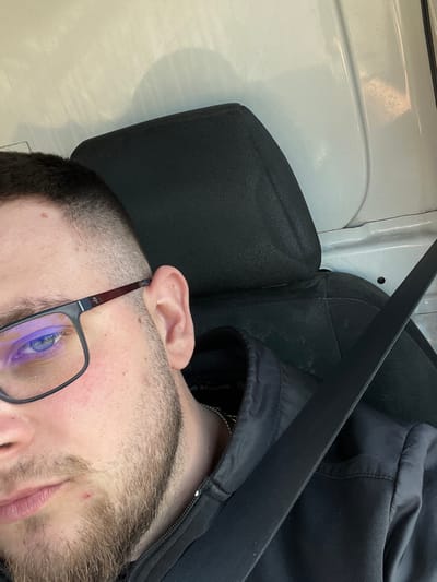 A male individual is depicted in a close-up, angled portrait within a vehicle in Koilovtsi, Bulgaria. The subject, visible from the forehead to the upper chest, has short, dark hair with a faded cut, light stubble, and blue eyes. He wears black-framed rectangular glasses, with a distinct purple-blue reflection on the left lens. A small mole is present above his left eyebrow, and a red blemish is visible on his chin. He is dressed in a black zip-up jacket, under which a silver-colored chain is visible around his neck. A black seatbelt extends diagonally across his chest and shoulder. Behind him is a black fabric headrest and seat back, consistent with a vehicle interior. The background features a light-colored, utilitarian paneling, suggesting the inside of a van or similar vehicle, with subtle textures and reflections from ambient lighting.