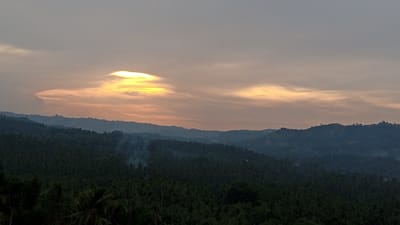 This photograph captures a serene landscape in Buketmulia, Indonesia, during what appears to be either sunrise or sunset. The sky is filled with a gradient of colors, ranging from a muted gray at the top to warm hues of orange and yellow closer to the horizon. A distinctive cloud formation, brightly illuminated by the sun, is visible in the upper left quadrant of the image.
Below the sky, a series of rolling hills covered in dense, dark green vegetation dominate the middle ground and foreground. The vegetation appears to be a mix of trees, possibly including palm trees, which are characteristic of tropical regions. Wisps of smoke are rising from several points within the forested hills, suggesting some form of controlled burning or a small, localized fire.
The overall impression is one of natural beauty and tranquility, with the warm light of the sky contrasting with the dark, lush landscape. There are no visible people or animals in the frame. The image appears to be a wide-angle shot, encompassing a vast expanse of the natural environment.