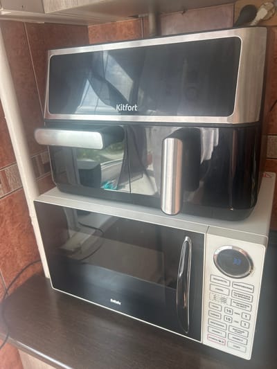 The image shows a stacked appliance setup on a dark brown countertop, likely in a kitchen, with a tiled backsplash. On top is a black and silver air fryer with the brand name "Kitfort" visible on its front. Below the air fryer is a silver microwave oven with the brand name "bbk" on its lower front panel. The microwave has a black glass door and a control panel with various buttons and a digital display showing "33". The scene is indoors, and the lighting suggests it could be daytime. The overall impression is of a functional kitchen appliance arrangement. The location of Kokhma, Russia, is a detail that cannot be determined from the image alone and is therefore not included in the description.