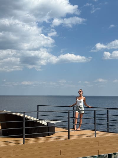 The image captures an outdoor scene on a bright day in Hlibivka, Ukraine, featuring a large body of water. A single female subject stands approximately center-right on a light-colored wooden or composite deck. She is a young adult, with brown hair styled in a bun, wearing sunglasses, a white tank top, and light-colored denim shorts. She stands barefoot, resting both hands on a dark gray metal railing that runs horizontally across the foreground, separating the deck from the water. To the left of the subject, a large, dark brown wicker-style circular lounge chair with light-colored cushioning is present on the deck. Beyond the railing, a vast expanse of calm, dark blue-gray water extends to the horizon, indicative of a large body such as the Kyiv Reservoir. The sky above is clear blue with scattered white cumulus clouds.