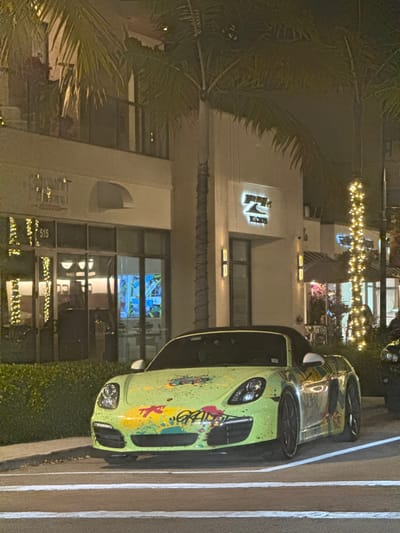 The image displays a custom-painted lime green Porsche Boxster convertible parked at night on a paved surface along a commercial street. The vehicle features a graffiti-style design with splashes of yellow, pink, and black paint, including the partial word "GRAFF" on the front bumper. Its black soft-top roof is lowered. In the background, well-lit, multi-story commercial buildings are visible. A prominent illuminated sign for "PURE YACHTS" is central, adjacent to another establishment marked with "515" and partial lettering "CHANT..." on its facade. Several palm trees, one adorned with string lights, are integrated into the streetscape. The scene is located on 5th Avenue South in Naples, Florida, United States. No individuals are clearly discernible or actively interacting within the visible frame.