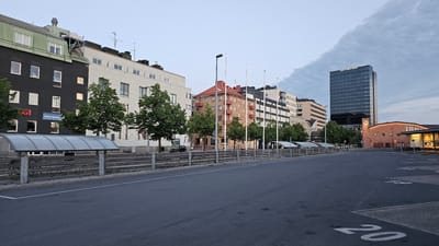 The image shows an outdoor view of a street and buildings in Jönköping, Sweden. In the foreground is a paved area with white markings on the ground, including the number "20". To the left, there is a sidewalk with a fence and covered bike racks. Behind the sidewalk are several multi-story buildings of various architectural styles. The buildings are residential and commercial, with different colored facades and window arrangements. Several trees are planted along the sidewalk. In the background, a tall, dark glass skyscraper stands out, with a sign that reads "QUALITY HOTEL MATCH". To the right of the skyscraper is a brick building with arched windows and a clock. The sky is overcast with some clouds. The scene appears to be captured during daylight, possibly in the early morning or late afternoon, given the soft lighting.