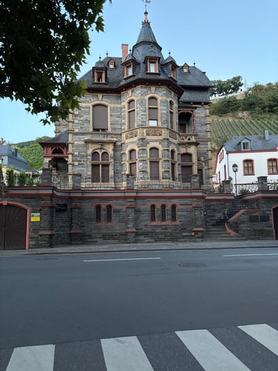 The image shows a multi-story building with a stone facade and dark grey roof, located in Bernkastel-Kues, Germany. The building features several architectural elements, including arched windows with dark shutters, decorative stonework, and turrets. A red trim accents the roofline and various sections of the facade. In front of the building is a paved street with a pedestrian crossing. To the right of the main building is a smaller white structure with red trim around its windows. The background reveals a steep hillside covered with vineyards. The sky is clear and blue.