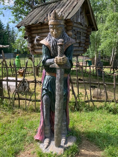 In the city of Pushkinskie Gory, Russia, a carved wooden statue of a king stands outdoors. The king is depicted wearing a crown, a long beard, and armor, and is holding a large sword in front of him. Behind the statue, a rustic wooden structure, resembling a small cabin or hut, is visible. The scene is set on a grassy area, with a wooden fence in the foreground and trees in the background. The overall impression is one of a whimsical, possibly historical, outdoor display.
