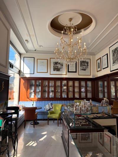 The image depicts the interior of a shop or cafe in Grad Opatija, Croatia. The establishment features a high ceiling with an ornate chandelier and decorative moldings. The walls are adorned with framed artwork. A tall, dark wood cabinet with glass doors displays various items. Glass display cases showcase confectionery items. A few chairs provide seating near the display cases. Sunlight streams through the windows. The overall ambiance is upscale and inviting.
