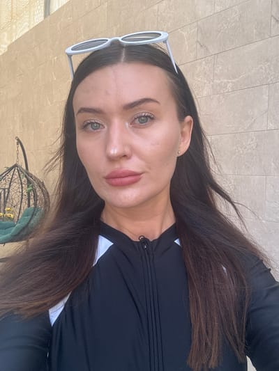 The media file shows a young woman, who is the primary subject, positioned in a selfie-style shot. She has long, dark brown hair, fair skin, and striking green eyes. She is wearing white-rimmed sunglasses and a black, zip-up top with white accents. The woman is looking directly at the camera, with a neutral or slightly inquisitive expression.
The background appears to be an outdoor setting with tiled flooring, suggesting a patio or public space. The lighting indicates it is daytime, and there are no immediate cues about the weather. In the lower part of the frame, a portion of a decorative metal basket is visible, possibly a hanging planter, with some green foliage inside.
The image appears to be taken casually, likely by the subject herself. The composition is close-up, focusing on the woman's face and upper body. The location context provided is Ayn al Basha, Jordan, which, combined with the tiled background and general aesthetics, could suggest a public area within a city or town. There is no visible text in the image.