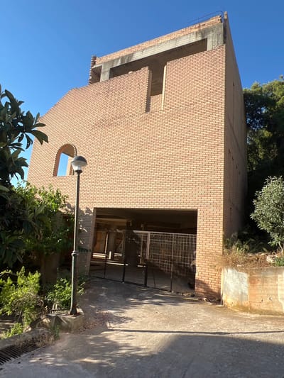 The image depicts an unfinished brick building with exposed rebar and concrete at the top, suggesting construction is ongoing or has been halted. The building is situated in an outdoor environment with a clear blue sky. To the left of the building, there are trees and lush green vegetation. A single lamppost stands in front of the building, and a driveway leads to a large opening with a metal gate, which appears to be an entrance to a garage or parking area. The ground is paved with concrete. The scene is located on a sunny day, casting shadows on the ground. The provided crops confirm the architectural details and surrounding vegetation.
