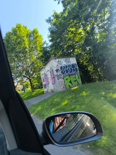 The image depicts a view from inside a car, likely moving, in an outdoor setting possibly in Saint-Pierre-de-Bœuf, France (though this is unconfirmed from visual data alone). The main subject is a small, concrete utility building or structure heavily covered in graffiti. The graffiti includes words like "REVS" and other illegible tags in various colors. The building is positioned on a grassy slope, near a paved pathway. Background details include trees with dense green foliage, bathed in sunlight. The sky is visible and is a clear blue. A car's side mirror is prominently visible, showing a blurred reflection of the car's interior and the passing scenery. The interior elements suggest that a person is in the driver's seat (indicated in the reflection). The scene appears to be taken during the daytime under sunny weather conditions.