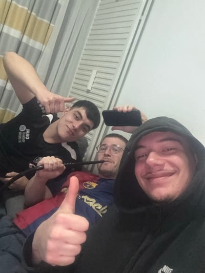 The image depicts three young adult males in an indoor setting, specifically identified as being in Krems an der Donau, Austria.
The leftmost male, positioned towards the rear, wears a black short-sleeved shirt bearing the text "JAKO TEAMSHOP BREITLER" and the number "89." He is smiling and extends his right hand with his index finger and thumb extended, forming a gesture.
The central male, seated between the other two, wears glasses and a blue and maroon short-sleeved jersey, indicative of FC Barcelona team colors. He is observed holding a black hookah hose to his mouth.
The rightmost male is in the foreground, wearing a black hooded sweatshirt with a white Nike swoosh logo on the chest. He is smiling broadly, looking directly towards the viewer, and offers a "thumbs up" gesture with his right hand. He holds a black smartphone in his left hand, positioned as if capturing a selfie of the group.
All three individuals exhibit smiling expressions. The background consists of a plain light-colored wall. To the left, a striped curtain or blind, displaying alternating grey and light beige/yellowish horizontal bands, is visible. A vertical white trim or slat runs alongside the curtain.