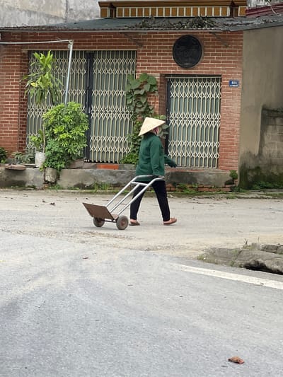 A person wearing a conical hat and a green jacket walks across a paved street in Hà Giang 2 Ward, Vietnam, pulling a two-wheeled handcart. The person is seen in profile, moving from left to right. The handcart is made of metal with a rusted platform and two spoked wheels. The background features a brick building with metal security gates and some greenery. A circular sign reading "Coffee" is affixed to the building wall. The street is partially wet and scattered with dry leaves.