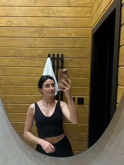 A young woman with dark hair, wearing a black tank top and black shorts, takes a mirror selfie in a bathroom. The bathroom walls are paneled with horizontal wooden planks. A white towel hangs on a black rack behind her. To her right, a black door frame is visible. On the wall to her right, there is a small digital display showing "25". The floor appears to be tiled. The woman is holding a smartphone with a pink case decorated with a Hello Kitty sticker in her right hand, and her left arm is resting at her side. She is wearing a silver necklace with a clear pendant and a thin silver bracelet on her left wrist. The overall setting suggests a modern, possibly minimalist, bathroom in a home or hotel. There is no specific information or visual cues in the image to indicate the location in the city of Stradch, Ukraine.