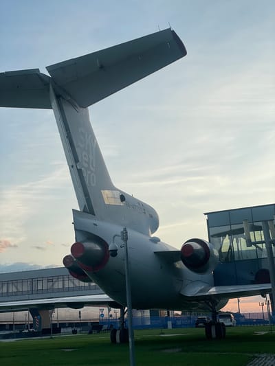 The image shows the tail section of a large, gray aircraft, possibly a Tupolev Tu-154, with its engines visible. The aircraft has text that appears to be "SKY JET PRO" on its tail fin, and a registration number that seems to read "RA-47379". It is parked on a grassy area, with a modern airport terminal building and walkway structure in the background. The sky is overcast with hints of sunset colors. The location is identified as Bol'shie Kabany, Russia. There are no people clearly visible in the image, though there may be a few figures sitting on benches in the distance. The scene appears to be an airport tarmac or an exhibition area at an airport.