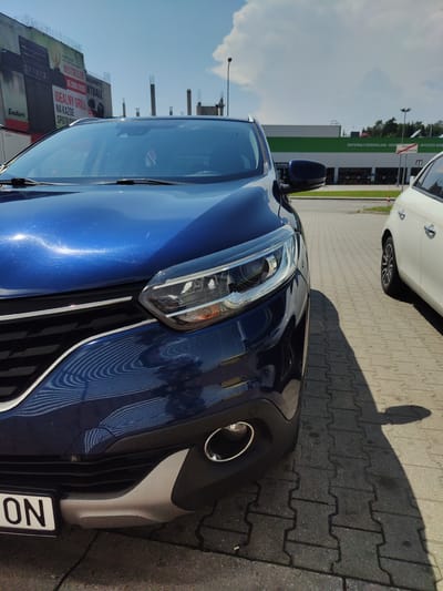 The image shows a dark blue car parked on a paved surface, possibly a parking lot, in Wieliczka, Poland. The car is positioned in the foreground, taking up the majority of the frame. Its front left side, including the headlight, grille, and part of the bumper, is clearly visible. The car's hood reflects the bright blue sky and scattered clouds, as well as some of the surrounding environment. A white car is parked to the right of the blue car.
In the background, a large building with a green and white facade can be seen, which appears to be a commercial establishment, possibly a hardware or building supply store, indicated by signage that reads "MATERIAŁY BUDOWLANE" (building materials) and "WYSTRÓJ WNĘTRZ" (interior design). Construction scaffolding and unfinished structures are visible beyond this building, suggesting ongoing development in the area. To the left of the frame, a large billboard displays advertisements for grills and other products.
The ground is paved with interlocking concrete blocks, typical of many outdoor public spaces in Poland. The lighting suggests it is daytime, with strong sunlight casting distinct shadows from the vehicles. There are no people visible in the image. The overall impression is a utilitarian, outdoor commercial setting on a sunny day.
