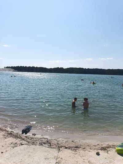 The image shows a sunny day at a lake in Yasnyska, Ukraine, with several people enjoying the water. In the foreground, two men are standing in the shallow water, facing each other. The man on the left has a beard and is looking down, while the man on the right is looking at the other man. The water is clear and calm, with some sunlight reflecting off the surface.

Further out in the lake, other individuals can be seen swimming, floating on inflatable tubes, and possibly kayaking. In the background, a dense forest borders the lake, and a large pile of white sand or gravel is visible on the left side. The sky is a clear blue with a few wispy clouds. The overall scene depicts a recreational area being utilized on a warm day.
