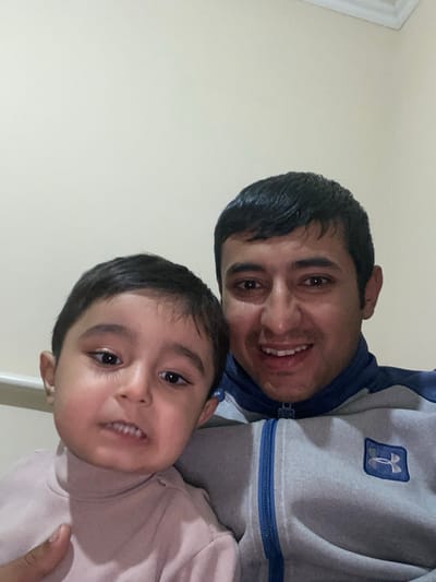 A close-up image taken in Galaosiyo, Uzbekistan, features two individuals. On the right, an adult male with short dark hair and dark eyes is visible from the chest up, smiling with teeth exposed. He wears a grey zippered jacket with blue accents on the collar, zipper line, and shoulders, featuring a dark blue square patch containing a white Under Armour logo on the right chest. To his left and slightly in front, a young child with dark hair and dark eyes is visible from the torso up. The child wears a light pink or beige long-sleeved turtleneck and has an ambiguous facial expression with a slightly open mouth, showing front teeth. The adult's left hand is partially visible, holding the child. Both individuals are positioned against a plain, light-colored wall, with a white molding visible in the upper right background.