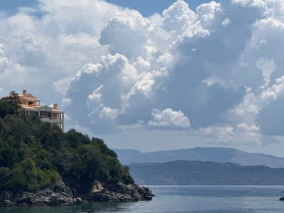The image depicts a coastal scene with a multi-story building situated on a vegetated hillside overlooking a body of water. The building, with its terracotta roof and white balconies, is positioned on the left side of the frame, atop a rocky and densely wooded incline. Lush green trees and shrubs cover the hillside. At the base of the hill, rough, dark rocks meet the calm, blue water. In the background, rolling hills and mountains recede into the distance under a sky filled with large, white cumulus clouds against a blue sky. The scene is tranquil and suggests a remote, possibly residential location. The specific location is in the Kassopaia Municipal Unit, Greece. There are no people or animals visible in the image.