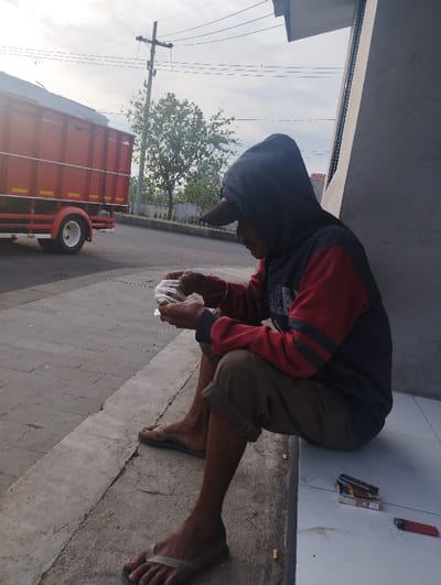 An individual is depicted sitting outdoors on a concrete ledge next to a tiled surface, likely a sidewalk or the entrance to a building in Kecamatan Jangkar, Indonesia. The person, appearing to be an older male, is in profile, looking down and focused on an item held in both hands, which is wrapped in clear plastic, possibly food. He is dressed in a dark hooded sweatshirt with red sleeves featuring black stripes, a dark baseball cap, light brown shorts, and olive green flip-flops.
Next to him on the tiled ground, a cigarette box (with colors hinting at a common Indonesian brand) and a red lighter are visible. In the background, a paved road stretches away, featuring a large red truck with white wheels, seemingly in motion. Beyond the road, a utility pole with overhead wires, green trees, and distant structures are present against a bright, slightly hazy sky.
The lighting suggests it is daytime, likely late morning or early afternoon, with soft, diffused sunlight. The scene conveys a moment of rest or a meal break taken by the roadside. The attire, the truck, and the general environment are consistent with a semi-urban or rural setting in Indonesia. No explicit text is clearly legible in the image.