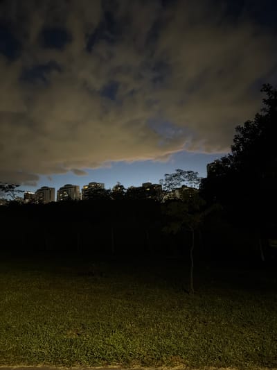 The image shows an evening scene in Brasília, Brazil. The sky is split with clouds and patches of blue, positioned to the left of the image. Several tall buildings with lit windows are visible between trees silhouetted against the sky. To the right of the buildings and trees is dark ground, transitioning into a patch of grass.