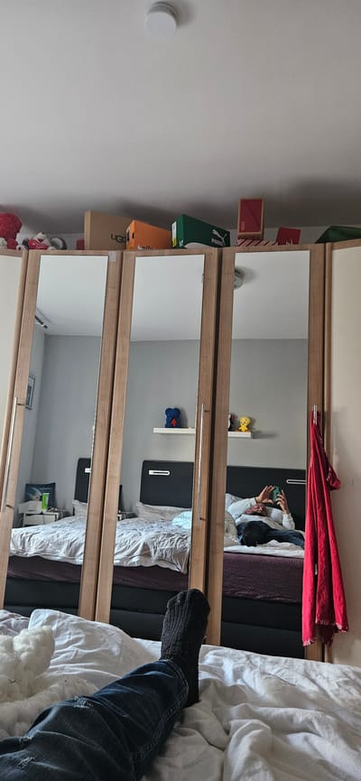 The image shows the interior of a room in Gütersloh, Germany. A person is lying on a bed, reflected in a mirrored wardrobe. The person is holding a phone, presumably taking the picture. The bed has a black headboard, patterned sheets, and white bedding. A leg wearing jeans and a black sock is visible in the foreground. The wardrobe has shelves on the left side holding items, including shoe boxes and decorative objects. A red cloth is draped on top of the wardrobe. A smoke detector is mounted on the ceiling.