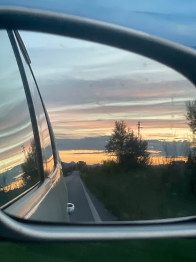The image captures a reflection in a car's side-view mirror, showing a road receding into the distance under a dramatic sunset sky. The sky is filled with horizontal bands of color, ranging from pale blue at the top to vibrant oranges and pinks closer to the horizon, interspersed with darker clouds. Trees and a solitary power line tower are silhouetted against the colorful sky. The mirror itself, with its silver trim, frames the scene. A sliver of the car's interior, including a white door handle, is visible on the right side of the mirror. The reflection also shows the side of the car and its movement along a paved road with a white line. The foreground outside the mirror shows a blurred green landscape, suggesting the car is in motion. There are no people or animals visible in the image. The scene appears to be a rural or suburban setting in Hvostiane, Bulgaria, during twilight.