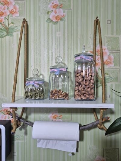 The image depicts a decorative shelf mounted on a wall covered with floral-patterned wallpaper. The shelf is made of white material and is suspended by thick ropes attached to the ceiling hooks. Three glass jars with decorative lace trim rest on the shelf, containing pumpkin seeds, walnuts, and cashews, respectively. Below the shelf, a roll of toilet paper is mounted on a branch. To the left of the toilet paper, a black and silver object, possibly an appliance, is partially visible. The scene is set indoors, in what appears to be a residential space. There is no indication of the specific location in Koçkor-Ata, Kyrgyzstan, within the image itself.