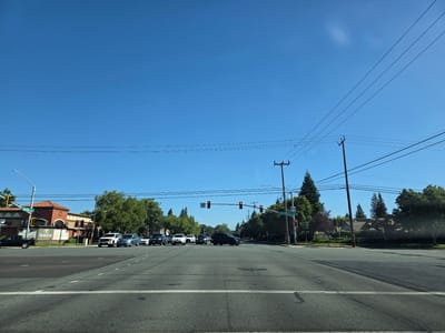 A street scene shows an intersection on a clear, sunny day. Several cars are stopped at a red light, waiting to proceed. A building with a red tile roof and arched windows is visible on the left side of the intersection, with a sign that reads "Laguna" above a logo. Other buildings and trees line the sides of the road. Power lines crisscross the sky, supported by wooden poles. A street sign indicates "Elk Grove Blvd" and an arrow pointing straight, suggesting the location is Elk Grove, California. The time of day appears to be late morning or early afternoon due to the bright sunlight and clear blue sky. No people are visible in the scene.