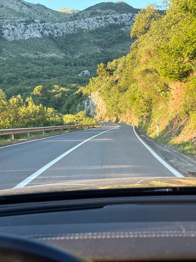 The image shows a scenic view of a winding road in Stupovi, Montenegro. The road is asphalt with white lines marking the lanes. A metal guardrail runs along the side of the road, separating it from a steep, tree-covered mountainside. The mountains in the background are rocky and sparsely vegetated at the top, transitioning to dense green foliage lower down. The sun is shining, casting long shadows and illuminating the trees with a warm, golden light. The perspective is from inside a vehicle, looking out through the windshield, with a portion of the dashboard visible in the foreground. There are no people or animals visible in the scene.
