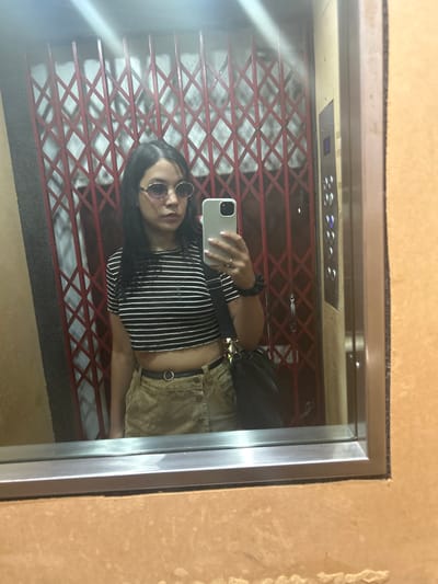 A person with dark hair and round sunglasses takes a mirror selfie in an elevator. They are wearing a striped crop top and tan shorts with a black belt. The elevator has a red lattice-style gate and a control panel with buttons. The mirror is framed with silver trim, and the surrounding walls appear to be beige.