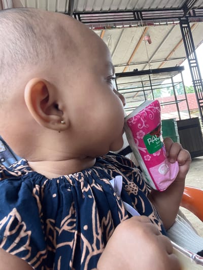 A close-up, profile view shows a young child, likely an infant or toddler, with a shaved head and dark brown eyes. The child is wearing a dark blue garment with a light brown pattern and a white ribbon at the neckline. The child is holding a pink and white packaged item, possibly a juice box or pouch, and appears to be drinking from it. The child has a small stud earring in their left ear. The background suggests an outdoor or semi-outdoor setting, possibly a patio or covered area, with a corrugated metal roof overhead supported by black metal beams. Some greenery and buildings are visible in the distance. The scene is well-lit, suggesting daytime. The environment appears to be a residential area in Seumirah, Indonesia.