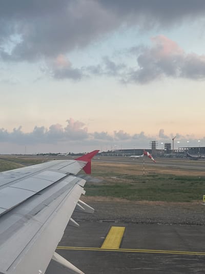 This image depicts an airport environment, viewed from the window seat of an aircraft, looking over its left wing. The scene is consistent with an international airport, such as Dalaman Airport in Dalaman, Turkey.

The foreground is dominated by the light grey wing of the aircraft, extending from the bottom left towards the center right of the frame. The wing shows operational wear and dirt, with visible panel lines and the red tip of the winglet on the far right. Below the wing, a dark asphalt taxiway is visible, marked with clear yellow lines: a solid double line and a thicker single line. Adjacent to the taxiway are areas of dry, yellowish-green grass.

In the mid-ground, a red and white striped windsock stands on a striped pole, showing a gentle inflation, indicating the presence of light wind. Beyond the immediate taxiway, further airport infrastructure is visible under a partially cloudy sky. Distant terminal buildings are illuminated, and a control tower structure is discernible. Several other aircraft are parked on the tarmac in the distance, one of which displays a partial Aeroflot logo on its tail. A large construction crane is also visible near some of the distant buildings, suggesting ongoing development.

The sky is filled with scattered clouds, predominantly grey, with some exhibiting soft pink and light blue hues, indicative of either sunrise or sunset conditions. No people are visible in the scene.