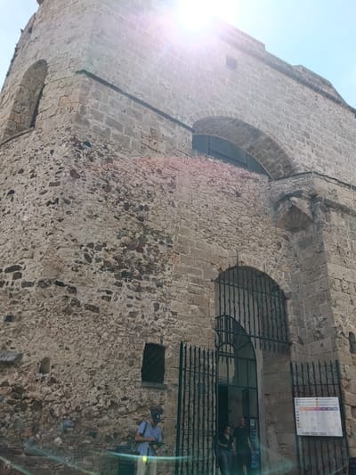 The image shows the exterior of a historic stone building, likely a church or fortress, in Alghero, Italy. The architecture features rough-hewn stone blocks and arched doorways and windows. Sunlight flares are visible in the upper portion of the image, indicating a bright day.
In the foreground, at the entrance of the building, a black wrought-iron gate is open, allowing access. Two individuals, a man and a woman, are standing just inside the gate. Further to the left, a person wearing a light blue shirt, beige pants, and a dark baseball cap is standing, looking down at something in their hands. They are carrying a bag over their shoulder.
A sign with multiple colored boxes and text in what appears to be Italian is mounted on the wall to the right of the entrance. The stone construction of the building is detailed, showing variations in stone size, color, and texture, with some areas appearing smoother and more intentionally worked, while others are rough and weathered. The overall impression is of an ancient and robust structure.