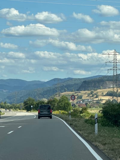 The image captures a scene on a highway, likely in the vicinity of Biberach, Germany, based on the signage and landscape. The perspective is from inside a vehicle, looking forward. A dark-colored minivan is driving in the right lane, ahead of the viewer. The road is paved asphalt with white lane markings. To the right of the road, there is a grassy verge with wildflowers and several white marker posts. A circular traffic sign with two red horizontal bars indicating a no-overtaking zone is visible.

The background features rolling hills covered in trees and fields, interspersed with houses of a small village. Several large electricity pylons with power lines stretch across the landscape. The sky is partly cloudy, with fluffy white clouds against a blue backdrop. The overall impression is a sunny day in a semi-rural setting.