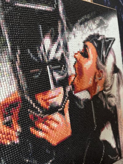 This media file captures a close-up, angled view of a diamond painting, an art piece created by adhering small, faceted resin beads to a canvas. The artwork features a stylized human figure, likely female, depicted in profile, facing towards the left. She has light-toned skin, rendered in various shades of orange and peach beads, and wears a dark headpiece or costume with prominent cat-ear-like features. Her hair appears light-colored, possibly white or very light gray, and is styled to the back.

The figure holds a firearm, possibly a rifle or gun, with both hands, suggesting an intense or focused activity. Her expression, though pixelated by the diamond beads, conveys determination or readiness. The background of the painting is predominantly dark, providing a stark contrast to the figure, with subtle hints of lighter shades and other colors visible in various areas. The image emphasizes the textured surface of the diamond painting, showcasing the individual reflective beads.

The overall scene portrays a character in action within the context of a mosaic art piece. No specific time of day, weather, or visible text can be discerned from the image. The location context provided for this image is Novyi Rizadei, Russia.