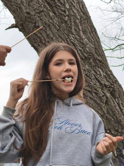 A young Caucasian woman is eating food off a stick, standing in front of a large tree. She has long brown hair and is wearing a gray hoodie with blue writing that says "Never Look Back". A hand holding a stick with food is visible to the left of the woman's head. The background shows a cloudy sky and other bare trees. It is difficult to confirm the location of the scene.