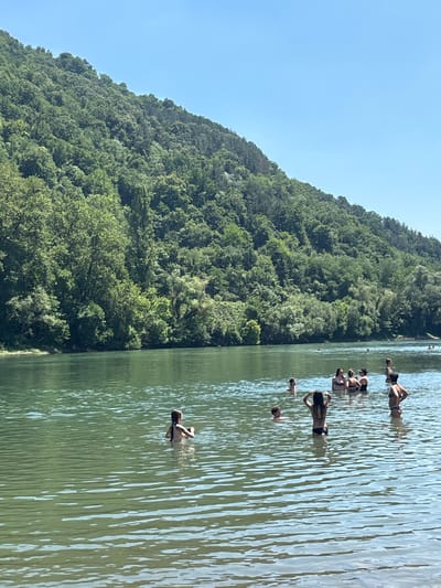 The image depicts a group of people swimming and enjoying themselves in a body of water, likely a river or lake. The scene is set against a backdrop of a densely forested, steep hillside. The water is calm with ripples reflecting the sunlight, suggesting a clear and bright day. The sky is a vivid blue, with no clouds visible, indicating pleasant weather. The time of day appears to be midday or early afternoon, given the strong sunlight.

Several individuals are visible in the water, some are standing and others are partially submerged. Their clothing appears to be swimwear, suggesting they are engaged in recreational swimming. The people are dispersed throughout the water, with no particular grouping or activity beyond general enjoyment of the water. The lush greenery of the hillside suggests a natural, possibly rural or semi-rural, environment. The presence of a forested hillside and a large body of water in Zvornik, Bosnia and Herzegovina, points to a location that is likely popular for outdoor recreation during warmer months. No specific objects other than the people themselves are prominent. There is no visible text in the image. The overall mood conveyed is one of relaxation and summer enjoyment.