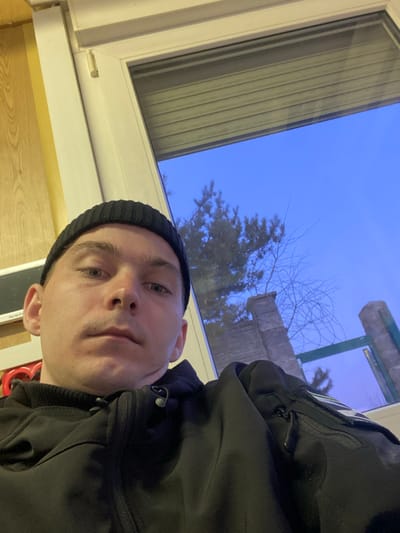 An image captures a close-up, chest-up view of a male individual indoors, situated in Марфино, Russia. The man, appearing to be in his 20s or 30s, wears a black knit beanie and a black zippered jacket with visible pockets and drawstrings. He looks directly forward with a neutral expression. Behind and to the right of the subject, a white-framed window reveals an exterior scene. The sky outside is a clear, deep blue, suggesting either dusk, dawn, or an overcast daytime. Visible through the window are dark green coniferous trees and bare deciduous tree branches. A light-colored stone pillar or structure is present in the middle ground, with a darker vertical element, possibly a fence post, further to the right. A partially raised beige roller blind is visible at the top of the window frame. To the left of the individual, an interior wall with light wooden paneling is partially visible, along with a rectangular silver object and a red object.