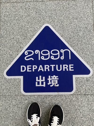 The image shows a blue arrow-shaped sign on a speckled gray tiled floor. The sign contains text in three languages. The top line displays text in a script that appears to be Lao. Below that, it reads "DEPARTURE" in English. The bottom line shows Chinese characters, "出境". Two black and white sneakers are visible at the bottom of the image, suggesting someone is standing on or near the sign. The context of the "Golden Triangle Special Economic Zone, Laos" indicates this is likely an airport or a border crossing point within that region. The overall scene is of an outdoor or indoor public space with directional signage.