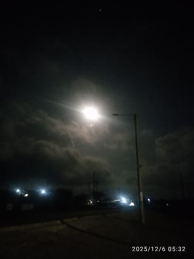 A night scene presents a brightly lit streetlamp, its luminescence piercing through the darkness and illuminating a road. Dark, looming clouds fill the sky, partially obscuring the moon, which appears as a bright, hazy disc. Dim lights from distant buildings punctuate the horizon, and what looks like a very thin crescent moon or bright star shines high above the clouds. The date "2025/12/6 05:32" is superimposed at the bottom right of the image.