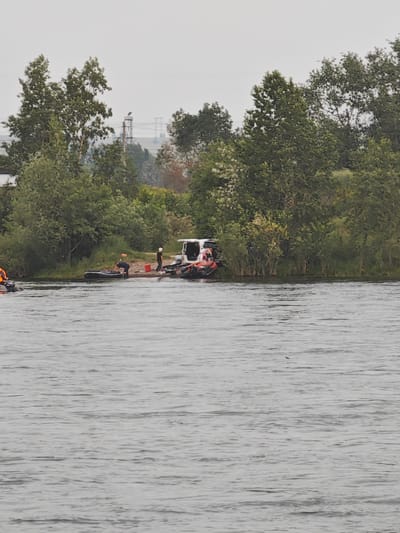 The image depicts a riverbank scene in Peschanka, Russia, with several individuals and watercraft gathered near the shore. The foreground is dominated by a wide body of greyish water with visible ripples. Along the riverbank, which is lined with lush green trees and some sparse vegetation, a white van is parked. Next to the van, on a sandy patch, a group of at least four people are present. One person, wearing a dark shirt, is bending over a deflated inflatable boat. Another person, in a black shirt and white cap, stands near the van. There are two inflatable boats partially visible on the water closer to the shore, one of which appears to have an orange object on it. Further out in the water, another small inflatable boat with at least one person wearing an orange life vest is visible. The background reveals more trees and a hazy sky. In the distance, a tall industrial tower with electrical wires is visible against the overcast sky, suggesting the presence of infrastructure beyond the immediate riverbank. The overall atmosphere suggests an outdoor activity or work being conducted by the river.