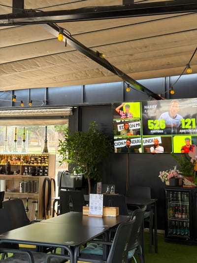 This media file depicts an outdoor seating area with a black metal pergola covered by a beige awning. A string of yellow lights is visible along the pergola. In the background, a large screen displays statistics and images related to "Harry Kane," with text indicating "526 PARTIDOS" and "121 ASISTENCIAS." Other panels on the screen show images of individuals, possibly soccer players, with text like "ED ON M" and "CIÓN MONDIAL UNDI."
To the right of the screen, a small glass-fronted refrigerator is stocked with bottled drinks. Further to the right, several black chairs are stacked. A small tree with green leaves and some pink and white flowers are also visible.
In the foreground, dark tables and chairs are arranged for dining or socializing. A shelf unit filled with various bottles is visible in the lower right portion of the image, suggesting a bar or restaurant setting. The scene appears to be during daylight hours, with natural light filtering through the awning and a window in the background that looks out onto a courtyard or garden area. The overall atmosphere suggests a relaxed, outdoor venue, possibly a sports bar or restaurant in La Vila Joiosa, Spain, given the context. There is no strong indication of a specific emotion conveyed by any visible subjects, and the weather appears to be pleasant.
