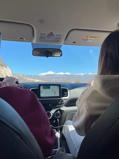 The image captures the interior of a Hyundai Venue vehicle from a rear passenger's perspective, traveling through a mountainous region in or near Miločani, Montenegro.

Two individuals occupy the front cabin. On the left, the driver is partially visible, with hands gripping the steering wheel, wearing a dark red/maroon long-sleeved garment. On the right, the front passenger is visible from the back of the head and shoulders, with long, dark hair, wearing a light-colored hooded sweatshirt and white trousers.

The vehicle's central infotainment screen displays a navigation application, with a blue route line indicating a current path and a remaining distance of approximately 9.7 km to a destination, identified by the Montenegrin text "Napred na sjever" (Go north) and "Kovačevića Ravan". Below the screen are circular climate controls and an automatic gear selector. A white charging cable is connected to the center console, extending towards the passenger's area. A brown paper-like bag rests on the floorboard on the passenger side.

Through the front windshield, a clear, bright blue sky is visible above a distant range of snow-capped mountains, illuminated by bright sunlight. A rugged, sparsely vegetated hillside appears on the immediate left of the vehicle's travel path. The rearview mirror is centrally positioned, and the right sun visor displays an 'AIRBAG' warning label.