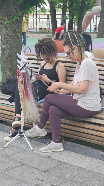 In the city of Alikonovka, Russia, two women are seated on a wooden park bench, engaged in hair braiding. The woman on the left, with dark dreadlocks, wears a black sports bra and dark leggings. She is actively working on the hair of the woman on the right, who has colorful dreadlocks and wears glasses, a light-colored t-shirt, and dark purple leggings. A tripod with attached hair extensions, some of which are pink and blonde, stands in front of the woman on the left. The environment appears to be a park or playground, with a sandy area and playground equipment visible in the background. A child in a red shirt is also visible in the background, near a large orange spherical object. The overall scene depicts a craft activity taking place in an outdoor public space.