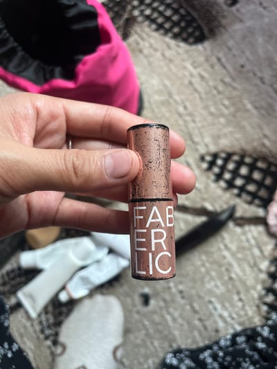 A close-up shot shows a hand holding a copper-colored lipstick tube with white lettering that reads "FABERLIC". The lipstick tube is positioned vertically, with the bottom half containing the text. The background is blurred and appears to be a textured surface, possibly a carpet or fabric, in neutral tones. There are out-of-focus objects in the background, including what looks like makeup tubes and a pink satin bag. The image was taken in the city of Ташлак, Kyrgyzstan.