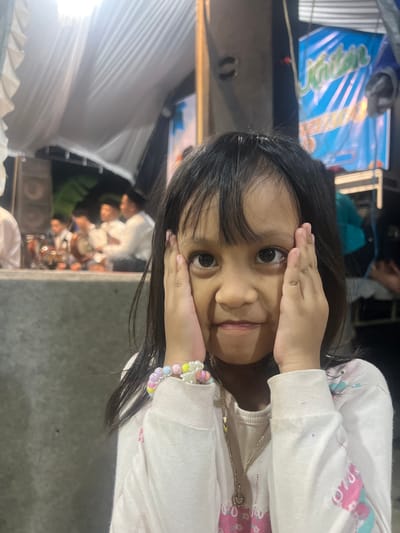In Wedarijaksa, Indonesia, a young girl with dark hair and dark eyes is captured in a close-up shot, her hands cupping her face as she looks directly at the camera with a playful expression. She wears a white long-sleeved shirt adorned with colorful floral patterns and a delicate beaded bracelet on her left wrist. A thin silver necklace hangs around her neck.

The background suggests an outdoor event. To the left, a stage area is visible with speakers and people dressed in traditional attire, some holding what appear to be musical instruments. A white canopy, possibly for shade or decoration, is stretched overhead. To the right, a blue banner with white and green text in a cursive font is partially visible. The lighting indicates it is likely evening. The overall setting seems to be a community gathering or performance in Wedarijaksa.