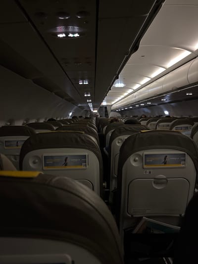The image captures the interior of an airplane cabin, focusing on a row of seats. The seats are upholstered in gray, with a headrest. The airplane's interior is visible along the left side, with lighting panels along the ceiling. The seats feature a card with "vueling CLUB avios" and the phrase "EVERY PURCHASE BRINGS YOU CLOSER TO YOUR NEXT TRIP." A safety notice about fastening seat belts is also visible on the headrests. People are seated further down the aisle, in the distance. The location is unknown, but it is known that Vueling Airlines serves Churriana, Spain.