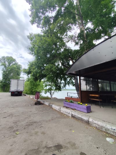 This media file shows an outdoor scene at a lakeside location in Raykov, Russia. The foreground is a gravelly, somewhat uneven ground, with a concrete curb running along the right side. A purple planter box with yellow flowers sits on top of the curb.
To the right, there is an open-sided covered structure, possibly a gazebo or picnic shelter, with a dark, translucent roof. Inside, wooden tables and benches are visible, suggesting a place for people to gather.
In the midground, a white box truck is parked on the gravel. Behind the truck and the covered structure, a large body of water, likely a lake or river, is visible, bordered by lush green trees. The trees are tall and dense, with a mix of broadleaf and possibly birch trees, judging by the distinct white bark on some trunks.
The sky is cloudy, with patches of white and gray, indicating an overcast or partly cloudy day. The overall impression is a slightly overcast day, possibly in the late morning or afternoon, in a natural setting by the water. There are no people visible in the scene, but the presence of the picnic shelter and tables suggests it is a place for recreation or rest. A small red metal object, possibly a trash bin or a signpost base, is situated near the curb between the truck and the shelter. The rear license plate of the truck has visible text, but it is too blurry to read.