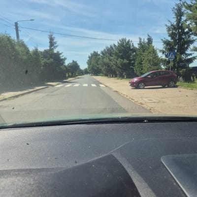 This is a photograph taken from inside a vehicle, looking out through the windshield. The image is oriented vertically, with the bottom of the frame showing the dashboard of the car. The scene outside depicts a road with a sidewalk on the left. A maroon car is parked on the sidewalk. Green trees line the sidewalk and the side of the road. A street lamp is visible on the left side of the image, along with power lines. The sky is bright blue with some wispy white clouds. There are some smudges and dust specks on the windshield. The lighting suggests it is daytime, likely a sunny day given the bright sky. A blue and white pedestrian crossing sign is visible near the parked car. The image appears to have been taken in Bralin, Poland. There is no visible text in the image.