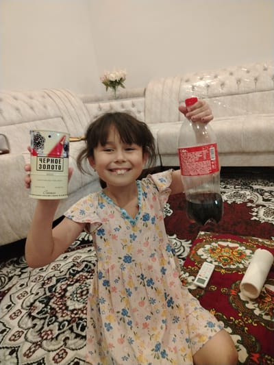 A young girl in a floral dress is seated on a carpeted floor, holding a can and a bottle of Coca-Cola. She is smiling at the camera. In the background, a sofa is visible. A remote control and a roll of paper are placed on the carpet near the girl. The scene appears to be an indoor setting, possibly a living room, in the city of Shovot, Uzbekistan.